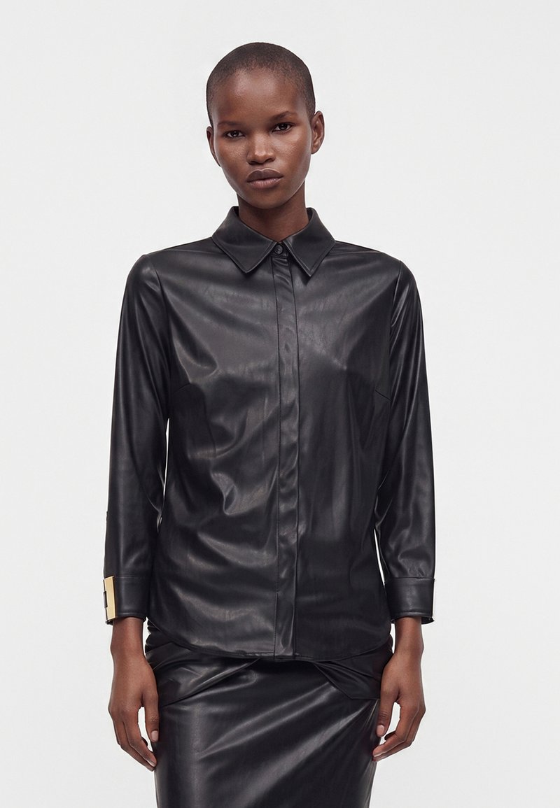 Femme aux cheveux courts portant une chemise en cuir noire boutonnée et une jupe en cuir assortie, debout devant un fond uni.