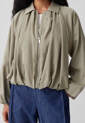 Femme portant une veste zippée ample de couleur olive claire avec des manches ballon et un jean bleu taille haute, main dans la poche, sur fond uni.