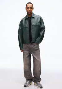 Ung mand stående iført grøn læderjakke med lynlås, sort skjorte, løse grå jeans og grå sneakers med hvide såler.