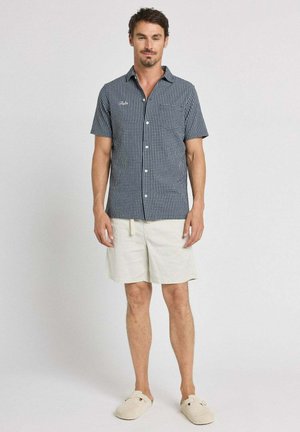 Homme debout portant une chemise à manches courtes à carreaux bleu marine, un short beige clair et des chaussures beiges sans lacets sur un fond clair uni.