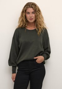 Maglione leggero di colore verde scuro con scollatura rotonda e spalle abbassate, abbinato a pantaloni neri aderenti. Design semplice con tessuto testurizzato.