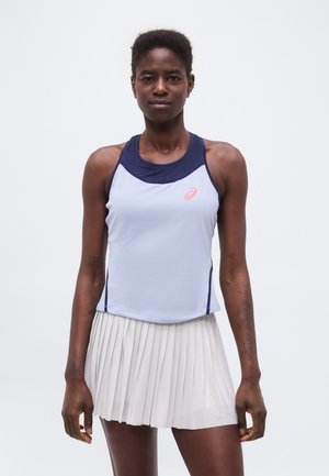Atlétikus nő áll, fehér és tengerészkék ujjatlan felsőt és fehér pliszírozott teniszszoknyát visel, egyszerű háttér előtt.