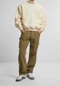 Cremefarbenes Fleece-Half-Zip-Pullover mit überschnittenen Schultern und gerippten Bündchen, kombiniert mit olivgrünen Cordhosen und weißen Sneakers mit schwarzen Akzenten.