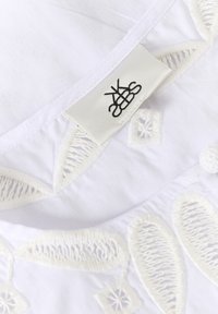 Tissu blanc avec des détails de dentelle complexes et une étiquette noire cousue portant un logo avec des lettres entrelacées.