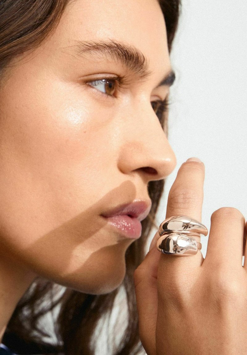 Sølvbelagt metalring med et slankt, buet design, der har to glatte, sammenflettede sektioner. Nærbillede af hånd og ansigtprofil.