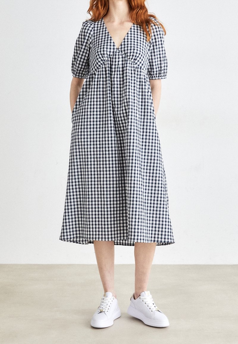 Femme en robe midi à carreaux noir et blanc avec manches bouffantes, debout, portant des baskets blanches sur un fond uni.