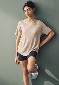 Top deportivo beige con rayas sutiles y un ajuste relajado, combinado con pantalones cortos negros y zapatillas de correr azul claro, frente a una pared verde.