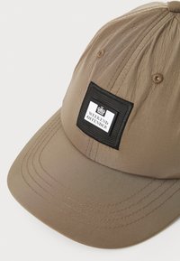 Beige baseball cap laget av lett stoff med buet skjerm, utstyrt med en svart firkantet lapp med teksten "WEEKEND OFFENDER" på fronten.