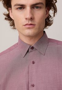 Giovane uomo dalla pelle chiara e capelli castano ondulati che indossa una camicia bordeaux con motivi e colletto a punta.