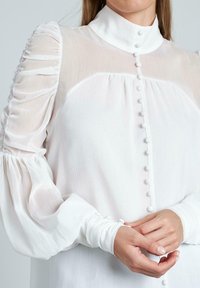 Blusa blanca de manga larga con cuello alto, con tela translúcida, mangas abullonadas y botones en el frente y en los puños.
