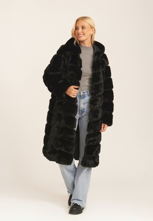 Black faux fur coat with a long, puffy design, featuring a shawl collar. Worn over a gray sweater and blue jeans, showcasing a casual look.