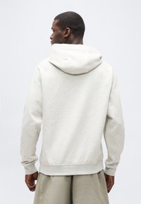 Άνδρας με ανοιχτ ό γκρι φούτερ με κουκούλα και μπεζ παντελόνι, γυρισμένος πλάτη, με φόντο ομοιόμορφο ανοιχτό γκρι.