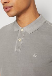 Homme portant un polo gris clair avec deux boutons en nacre et un petit logo brodé "M OP" sur le côté gauche de la poitrine.