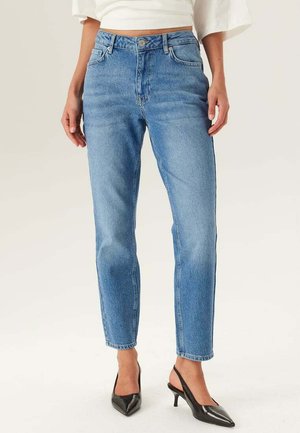 Kvinde iført lyseblå high-waist denimjeans og sorte spidse slingback-hæle, stående mod en ensfarvet baggrund.