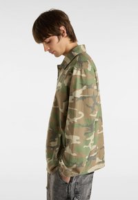Tarnjacke in grünen und braunen Farbtönen. Verfügt über einen Kragen, lange Ärmel und seitliche Taschen, aus einem leichten Stoff mit einer lockeren Passform.
