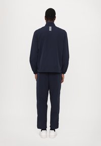 Tuta sportiva navy con polsini a coste e colletto alto. Presenta un dettaglio a righe verticali sulla schiena, abbinato a sneakers bianche.