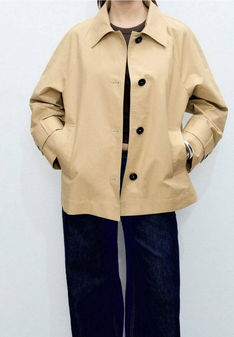 Personne portant une veste beige boutonnée avec les mains dans les poches, associée à un jean bleu foncé et un haut sombre en dessous.