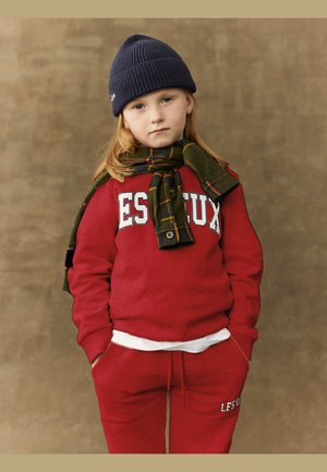 Sweat à capuche rouge avec le logo "ESTUX", pantalon de survêtement assorti, t-shirt blanc, écharpe en tartan vert et bonnet en tricot bleu marine. Texture lisse.