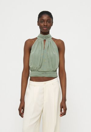 Žena v bezrukávovom, šalviovo zelenom crop hore so stojatým golierom a vysokými bielymi nohavicami, stojaca pred jednoduchým svetlým pozadím.