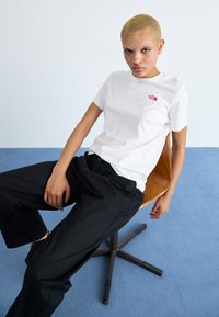 Hvit kortermet t-skjorte med rosa logo og avslappede svarte bukser. Modellen sitter på en tremøbel mot en lys blå bakgrunn.