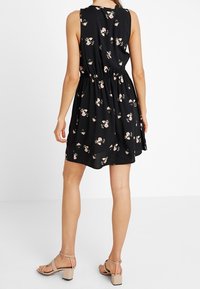 Kvinna i en ärmlös svart klänning med rosa blommönster, markerad midja och beige blockklackade sandaler, stående mot en enfärgad bakgrund.