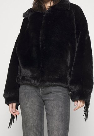 Persoon draagt een zwarte faux fur jas met brede mouwen en franjes, gecombineerd met grijze spijkerbroek tegen een effen achtergrond.