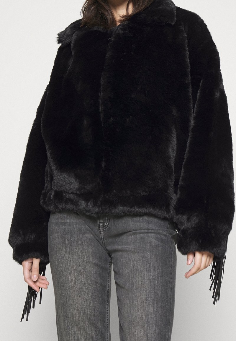 Personne portant une veste en fausse fourrure noire avec des manches larges et des détails en franges, associée à un jean gris sur un fond uni.