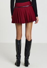 Donna che indossa una mini gonna rosso a plissettatura con una sottile cintura nera, un top a maniche lunghe nero e stivali in pelle nera fino al ginocchio, in piedi su un pavimento bianco.