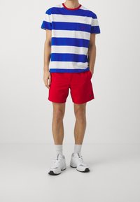 Blå och vit randig t-shirt med röd krage och liten bröstficka, i kombination med röda shorts. Vita sneakers och ankellånga strumpor.