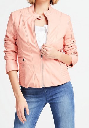 Personne portant une veste zippée rose clair sur un haut blanc et un jean bleu, ajustant la fermeture éclair de la veste d'une main.