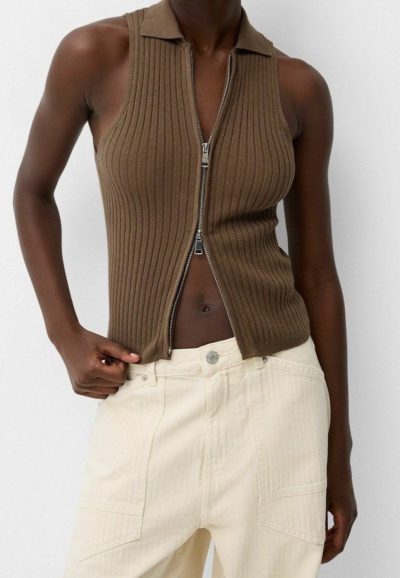 Personne à la peau foncée portant un haut sans manches en maille marron zippé et un pantalon taille haute couleur crème clair sur un fond blanc uni.