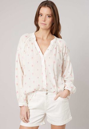 Vrouw draagt een losse witte blouse met roze patronen en witte shorts met opgerolde zoom, staand met één hand in de zak tegen een effen achtergrond.