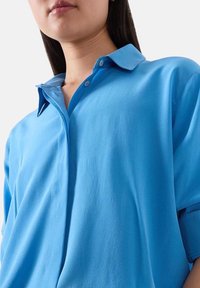 Camicia azzurra a maniche corte con colletto e dettagli con bottoni. Realizzata in tessuto morbido, presenta una vestibilità comoda e una leggera lucentezza.