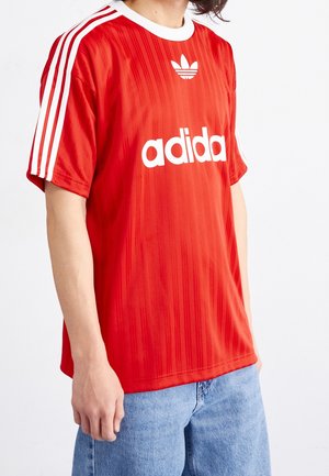 Rood Adidas sportshirt met witte strepen op de mouwen, een ronde hals en een groot wit logo over de borst; met verticale textuur.
