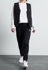 Svart väst med flera fickor över en vit långärmad skjorta, matchad med svarta sweatpants och vita Adidas sneakers med svarta detaljer.