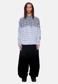 Νεαρός με μακριά ντραντλοκς φοράει μπλε και λευκό πουκάμισο καρό με μαύρα λουλουδάτα σχέδια στα μανίκια και φαρδιά μαύρα παντελόνια, στέκεται μπροστά.