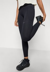 Leggings de sport noirs avec une texture lisse, présentant une petite poche latérale et des détails de couture contrastés. Portés avec des chaussures noires.