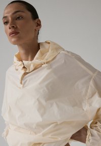 Kobieta z slicked-back włosami, ubrana w luźną, jasnobeżową kurtkę przeciwdeszczową z kapturem, patrząca nieco na bok na jednolitym tle.