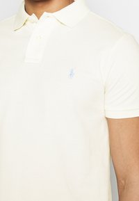 Polo tričko ve světle žluté barvě, vyroben é z texturované tkaniny. Obsahuje dvoubotkovou légu a malé modré logo na levé straně hrudi. Krátké rukávy.