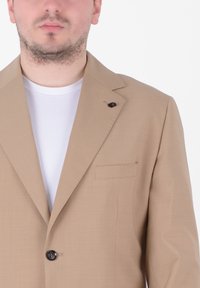 Blazer beige in tessuto leggero, con rever a lancia, chiusura con un solo bottone e tasca frontale. Indossato su una t-shirt bianca.