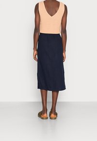 Béžový pletený top bez rukávů s V-zadním výstřihem, oblečený s sukní po kolena v námořnické modré barvě s bočním rozparkem, nošený s hnědými sandály na klínu.