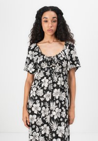 Sort blomsterkjole med hvide hibiscus- og bladmønstre, korte puffærmer og en firkantet halsudskæring med detalje i det samlede bryst.