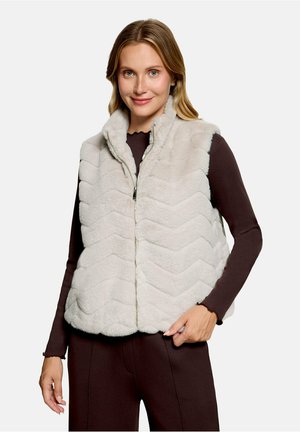 Femme portant une veste polaire matelassée gris clair par-dessus un haut à manches longues foncé, souriant avec une main reposant près de sa taille.