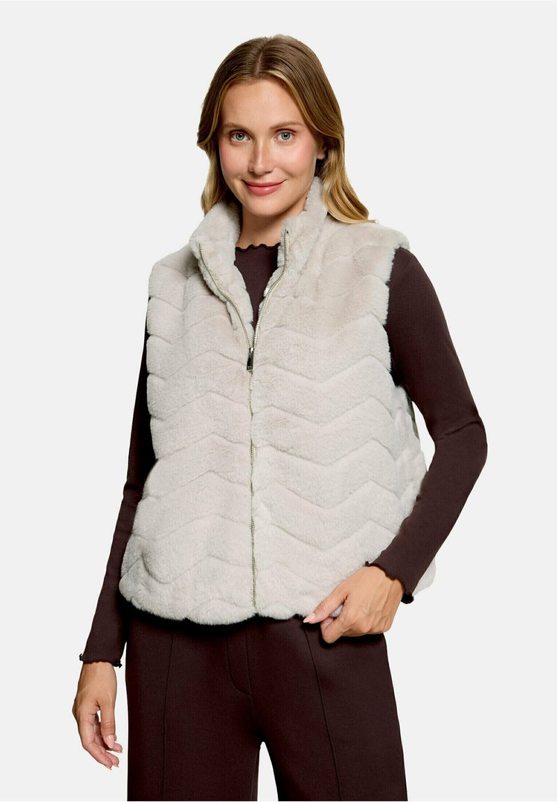 Femme portant une veste polaire matelassée gris clair par-dessus un haut à manches longues foncé, souriant avec une main reposant près de sa taille.