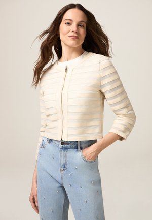 Veste en crème coupée avec des rayures horizontales, encolure ronde et fermeture éclair à l'avant. La mannequin porte un jean bleu clair avec des clous en argent dispersés.