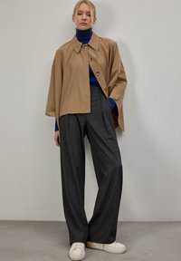 Femme posant dans une cape beige boutonnée, un col roulé bleu, un pantalon gris à jambes larges et des baskets blanches devant un mur uni.