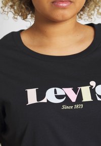 Persoon in een zwart shirt met een multicolore "Levi's" logo en de tekst "Sinds 1873" zichtbaar op de borst.