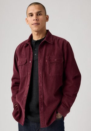 Chemise en velours côtelé bordeaux avec deux poches poitrine, boutonnage à l'avant et col. Portée par-dessus un t-shirt henley noir. Présente un tissu texturé.
