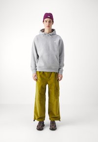 Grauer Kapuzenpullover, gelbe Cordhosen im Cargo-Stil und braune Schuhe mit Camouflage-Muster; ausgestattet mit einer Beanie mit Logo in Burgunderrot.