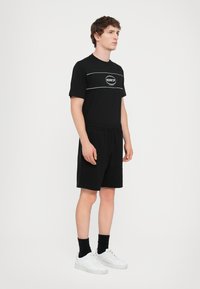 Black short-sleeve t-shirt with a logo on the chest, paired with black shorts. White sneakers and black socks complete the outfit.